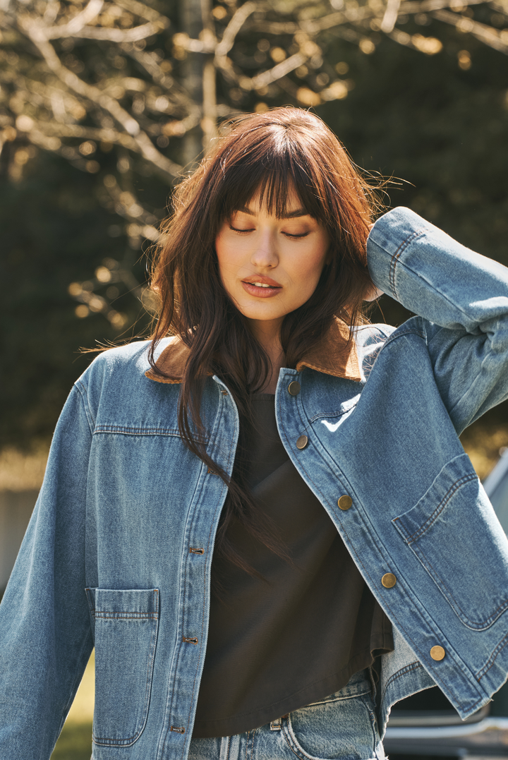 A woman wearing a light blue denim jacket with a brown corduroy collar over a black top, photographed outdoors with blurred trees in the background.