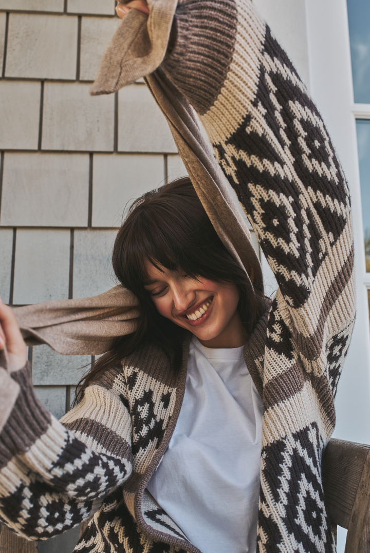 A woman smiling while wearing a chunky knit cardigan with a brown, beige, and black geometric pattern, holding the hood up over her head against a light gray shingled wall.