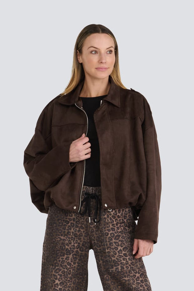 A model stands against a light gray studio background wearing a dark brown suede jacket, wide-leg brown leopard-print trousers, a black top, and black boots.