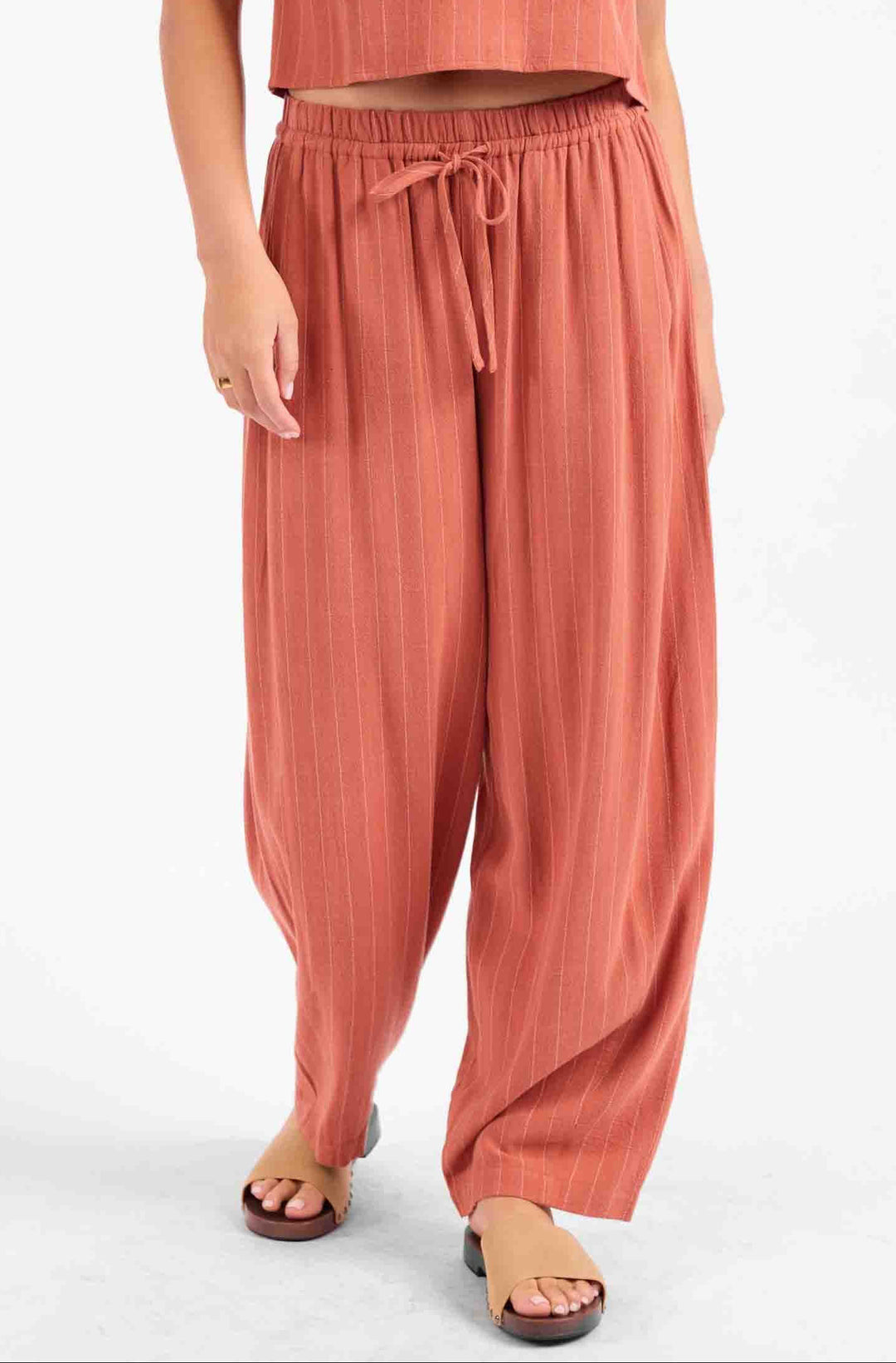 A model wearing a matching terracotta-colored two-piece set consisting of a short-sleeve cropped top and high-waisted, wide-leg drawstring pants, paired with brown slide sandals against a plain white background.