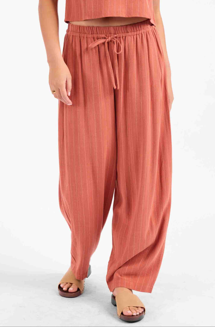 A model wearing a matching terracotta-colored two-piece set consisting of a short-sleeve cropped top and high-waisted, wide-leg drawstring pants, paired with brown slide sandals against a plain white background.