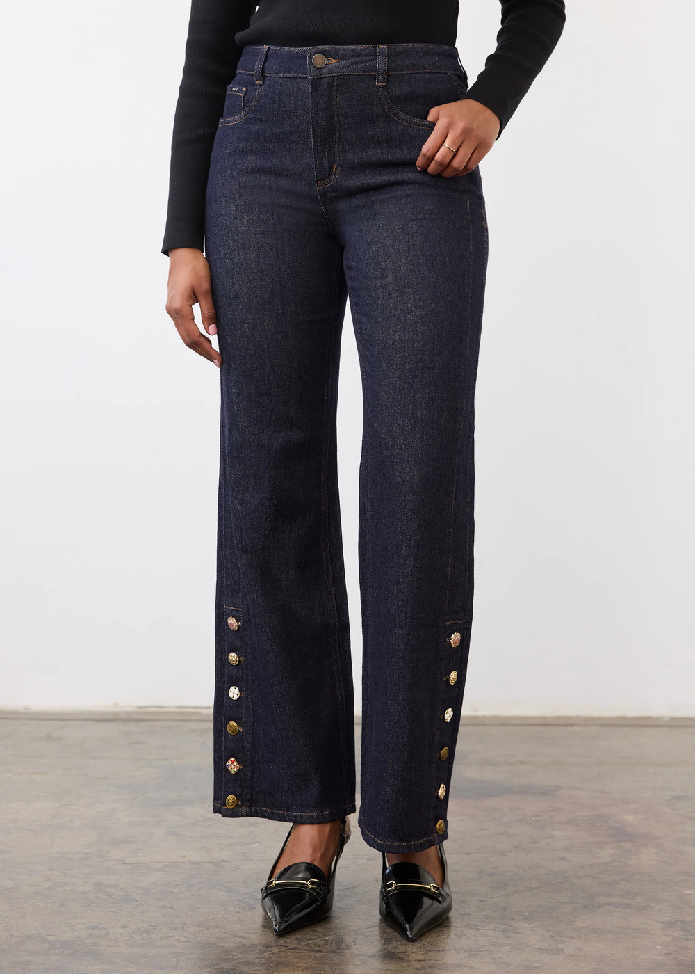 A model is shown from the waist down wearing dark blue flare jeans with decorative gold-tone buttons down the outer sides, paired with a black long-sleeve top and black pointed-toe flats.