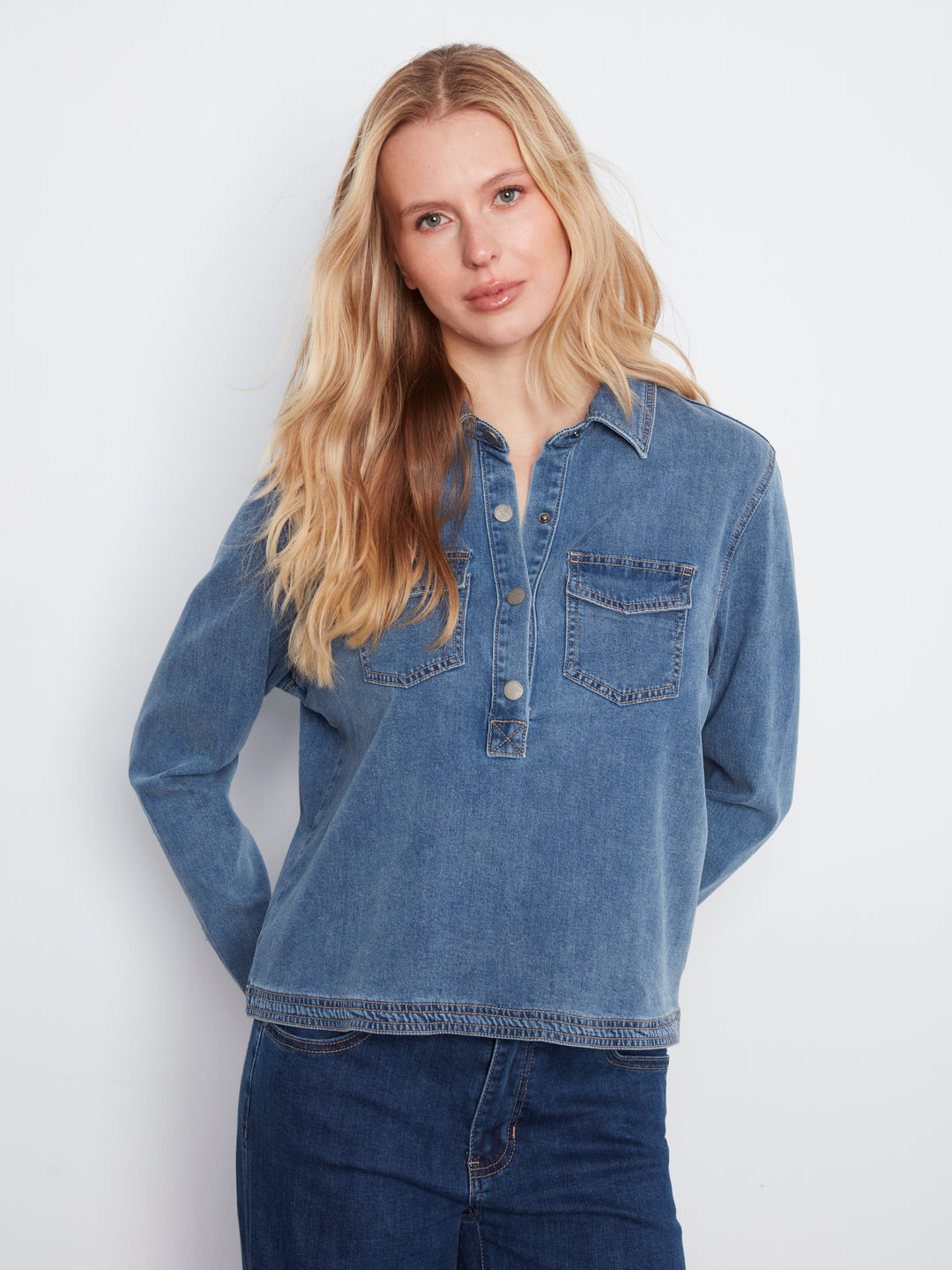 A blonde woman wearing a medium-wash blue denim long-sleeve button-up shirt with two chest pockets, paired with dark blue jeans, against a plain light gray background.