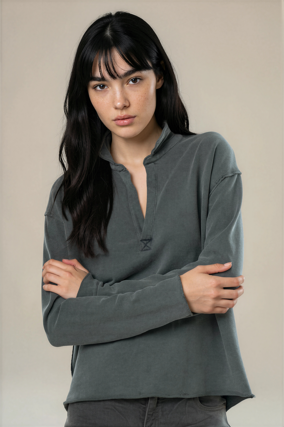 A woman with long dark hair wearing a dark gray long-sleeve henley shirt with a split collar and a small geometric logo on the chest, posed against a solid beige background.