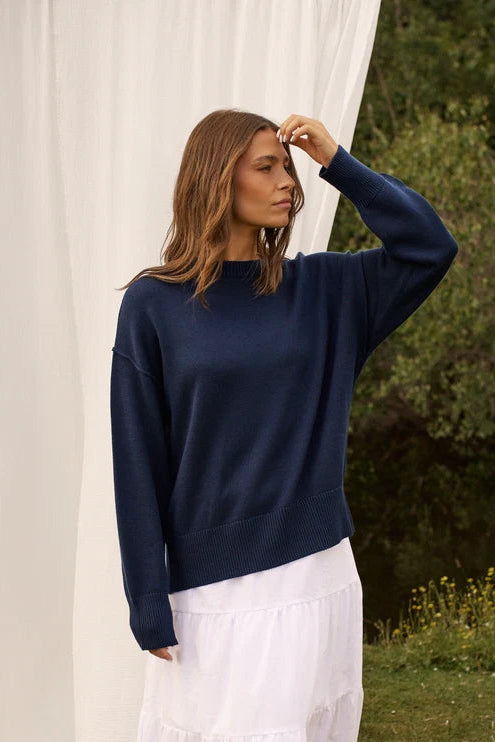 A woman stands outdoors wearing a navy blue long-sleeve crewneck sweater and a long white tiered skirt, with brown boots. A large white draped fabric backdrop is behind her, with grass and trees in the background.