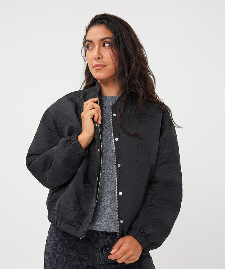 A woman modeling a black quilted bomber jacket with silver snap buttons, worn over a gray knit top and dark paisley-print pants, against a plain light gray studio background.