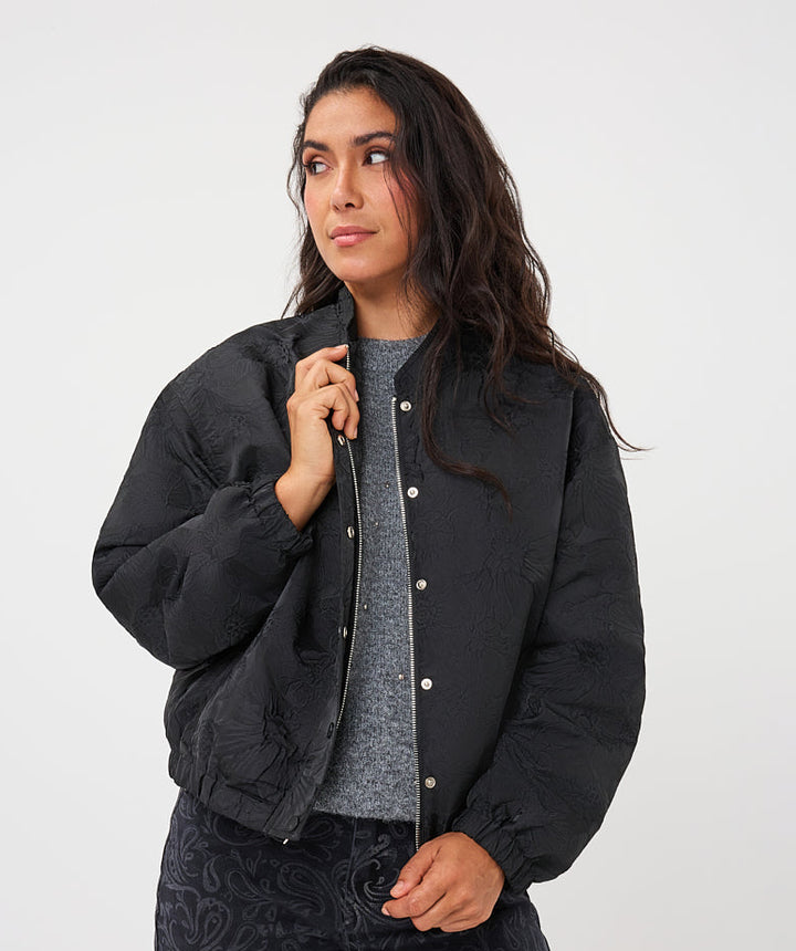 A woman modeling a black quilted bomber jacket with silver snap buttons, worn over a gray knit top and dark paisley-print pants, against a plain light gray studio background.