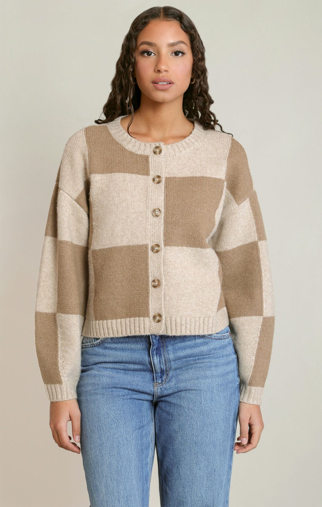 A woman wearing a beige and cream checkered button-up cardigan sweater with blue jeans, standing against a neutral light gray background.