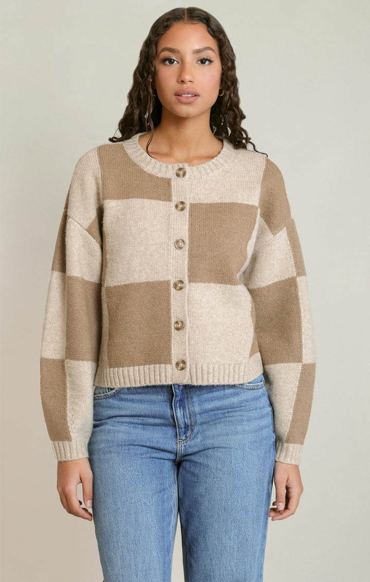 A woman wearing a beige and cream checkered button-up cardigan sweater with blue jeans, standing against a neutral light gray background.