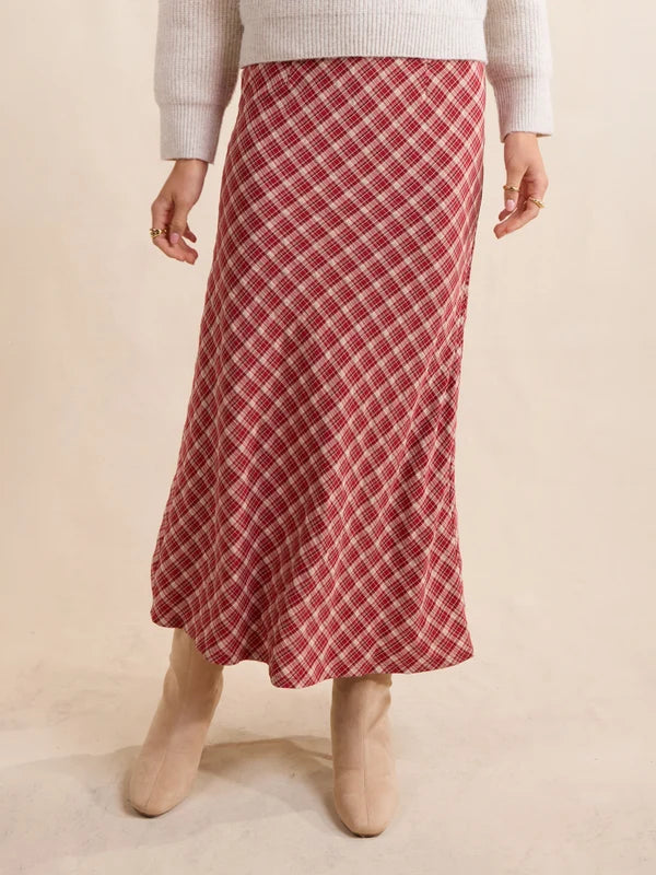 A red and white plaid midi skirt worn by a model, styled with a cream ribbed knit sweater and beige ankle boots against a warm beige studio background.