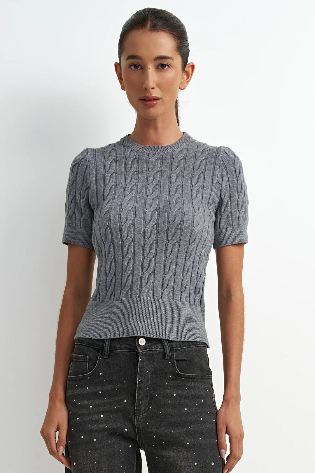 Woman wearing a gray cable knit sweater and black pants against a white background
