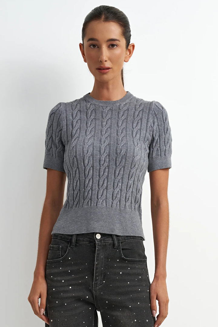 Woman wearing a gray cable knit sweater and black pants against a white background