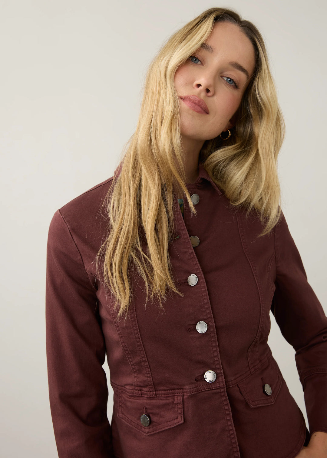 A woman with long blonde hair wearing a maroon button-up jacket with silver buttons, photographed against a plain light gray background.