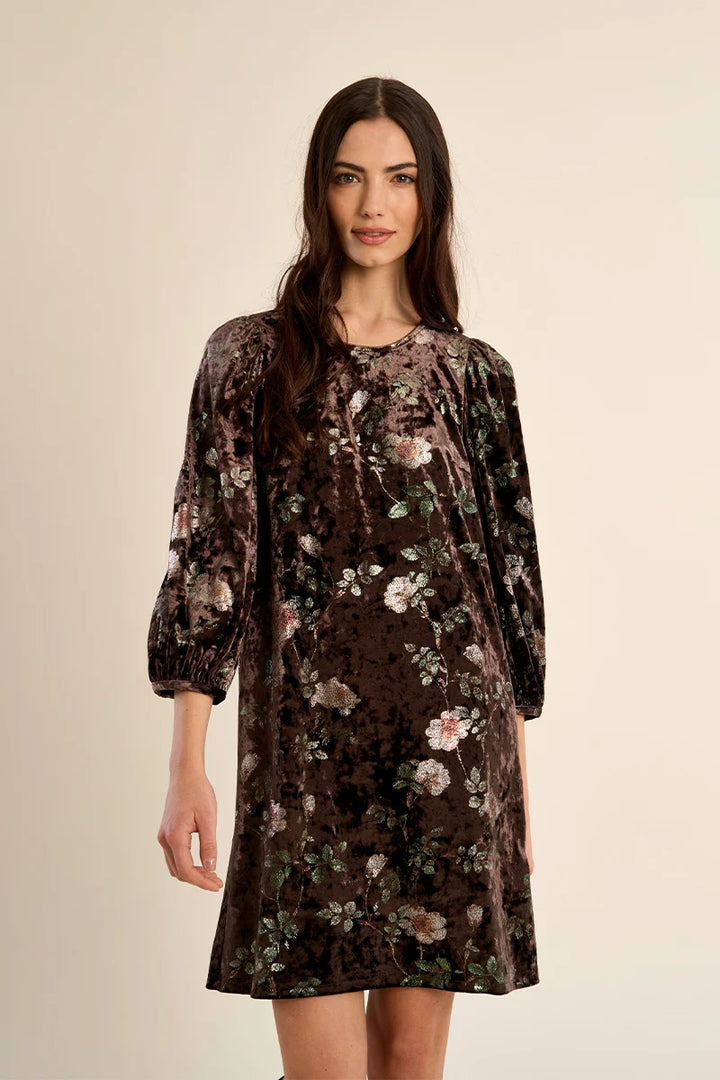 A woman modeling a dark brown velvet dress with a muted floral pattern in pink, green, and beige, set against a plain beige studio background.