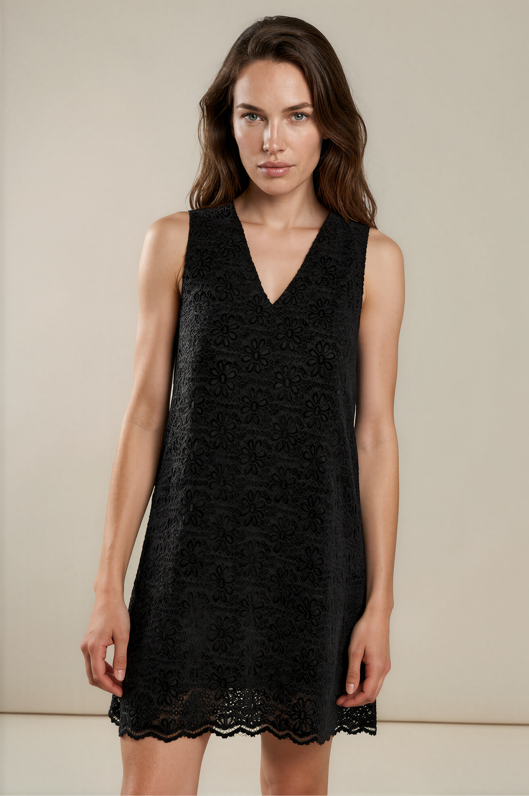A woman modeling a sleeveless black lace mini dress with a V-neckline against a plain beige studio background.
