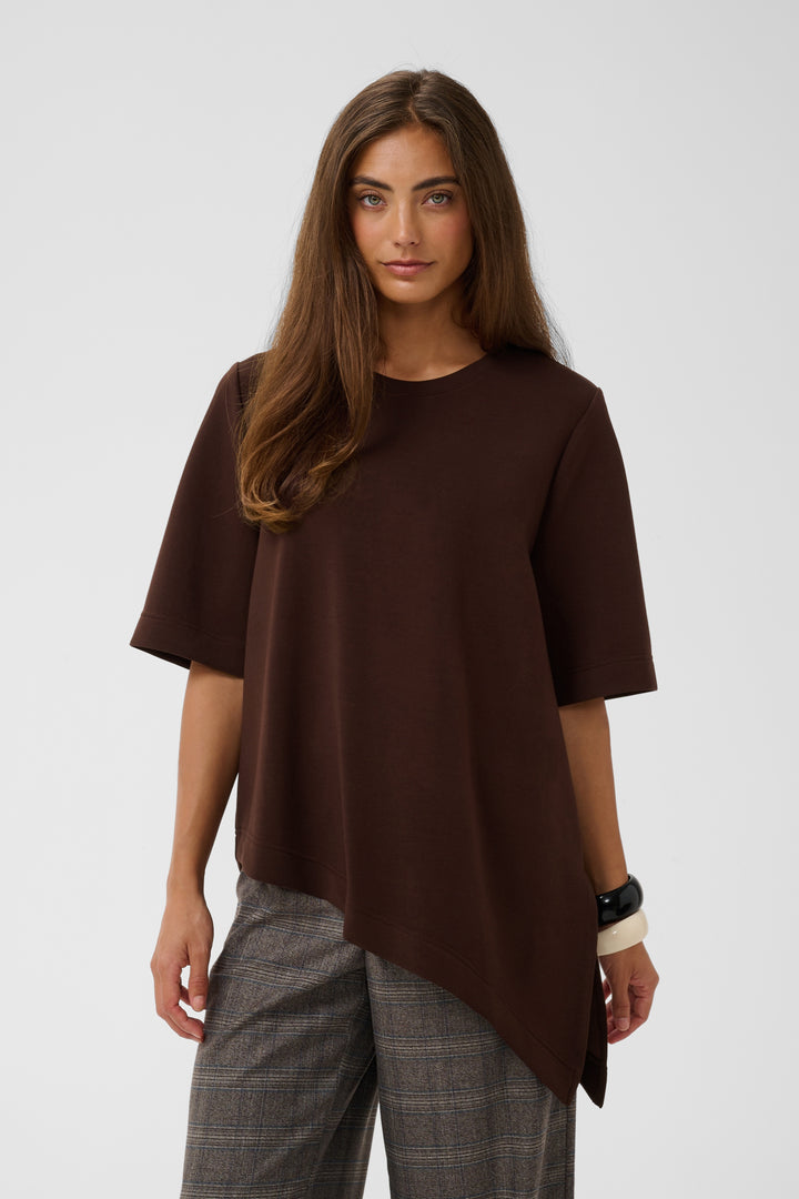 A woman modeling a dark brown short-sleeve top with an asymmetrical hem, paired with gray plaid trousers, against a light gray studio background.