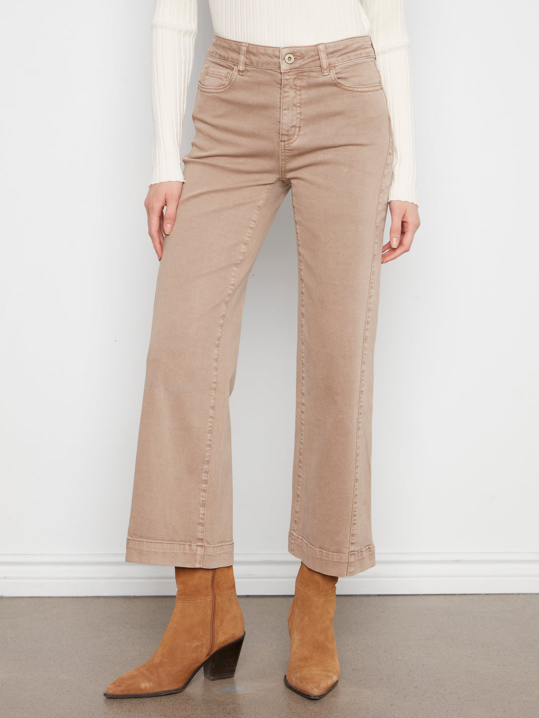 A model wearing beige cropped flare jeans, a white ribbed long-sleeve top, and tan ankle boots, standing against a white wall.