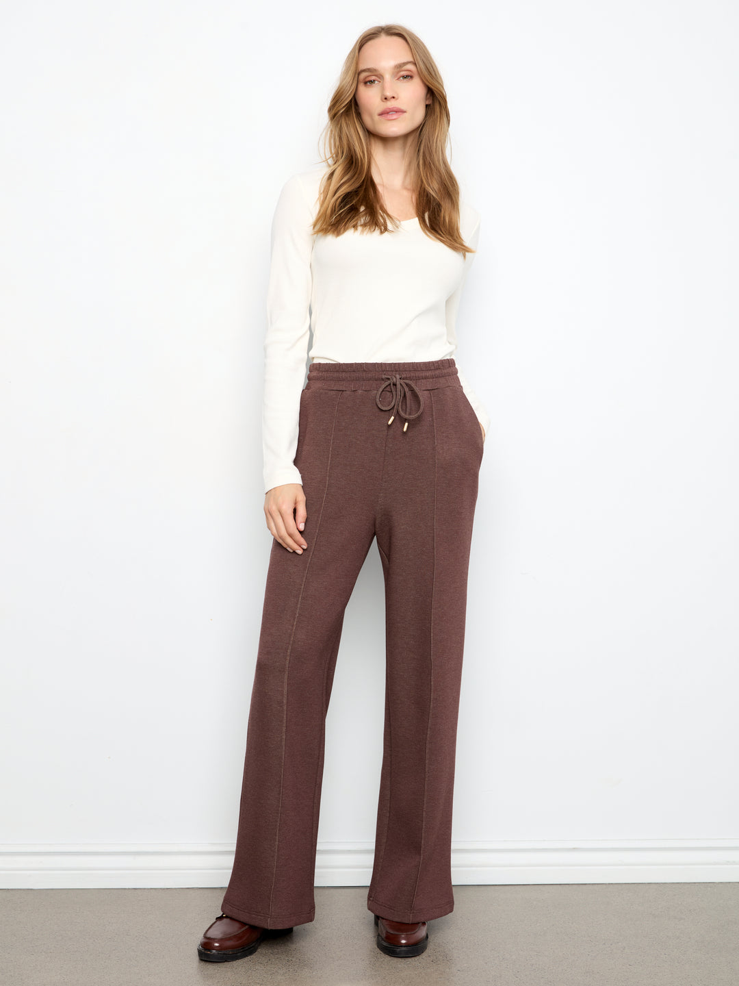A woman modeling a pair of wide-leg brown pants with a drawstring waist, paired with a white long-sleeve top and brown shoes, standing against a white wall.