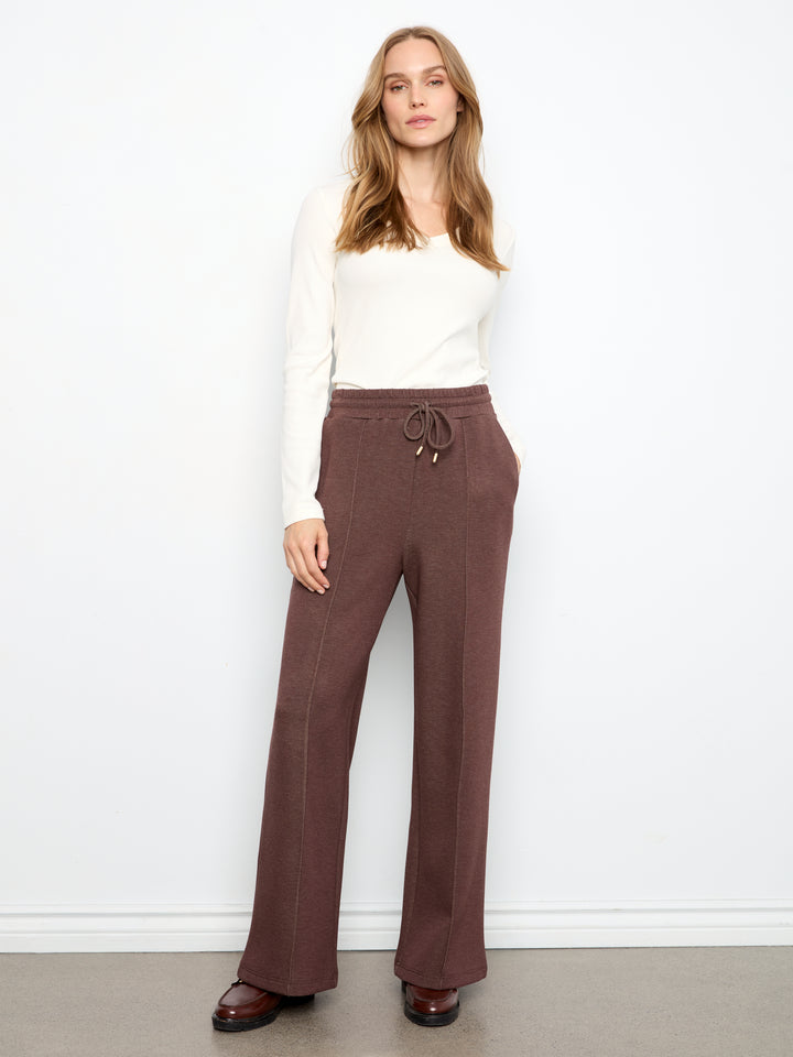 A woman modeling a pair of wide-leg brown pants with a drawstring waist, paired with a white long-sleeve top and brown shoes, standing against a white wall.