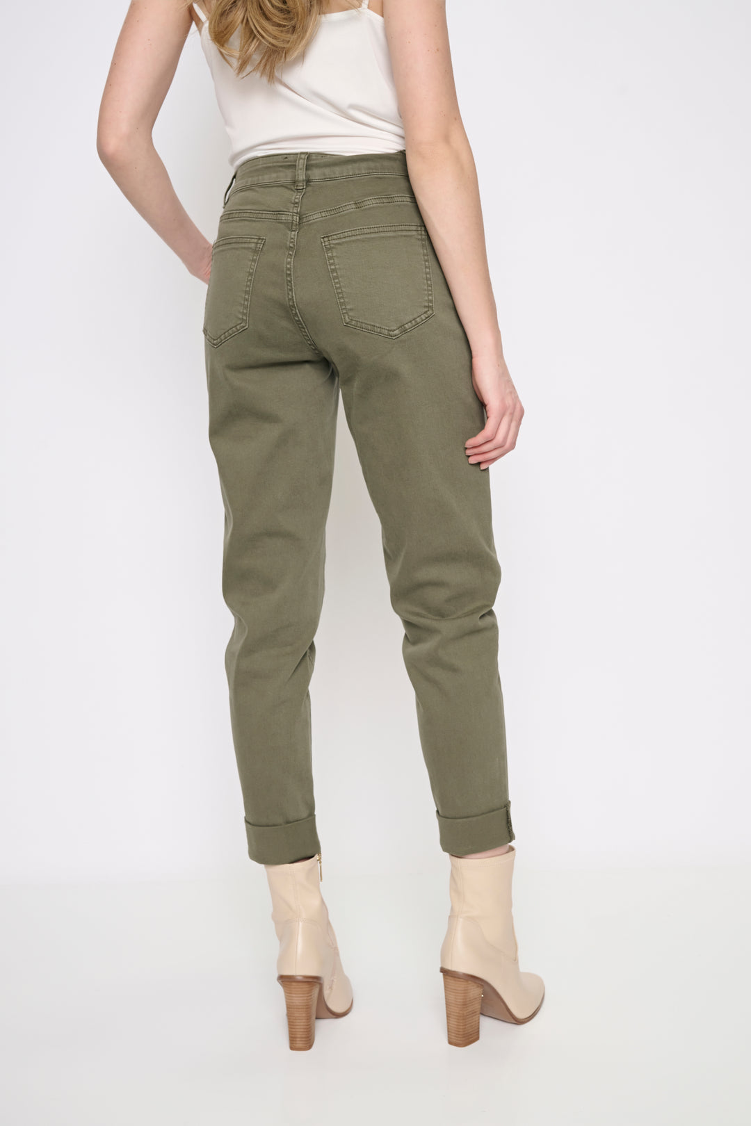 A woman shown from the back wearing olive green slim-fit jeans, a white sleeveless top, and beige ankle boots with wooden heels, against a plain white studio background.