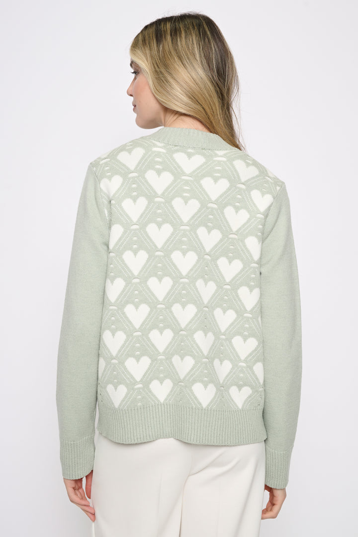 A woman shown from the back wearing a sage green knit cardigan with a white geometric heart pattern, paired with white pants, against a plain light gray background.