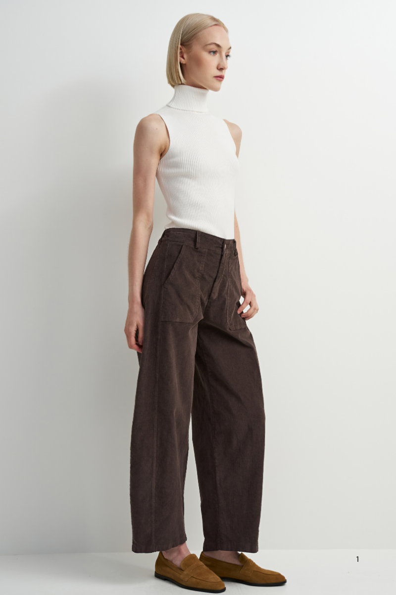 A model is shown from the side wearing a white sleeveless mock-neck top, dark brown wide-leg trousers, and brown suede loafers against a plain white studio background.