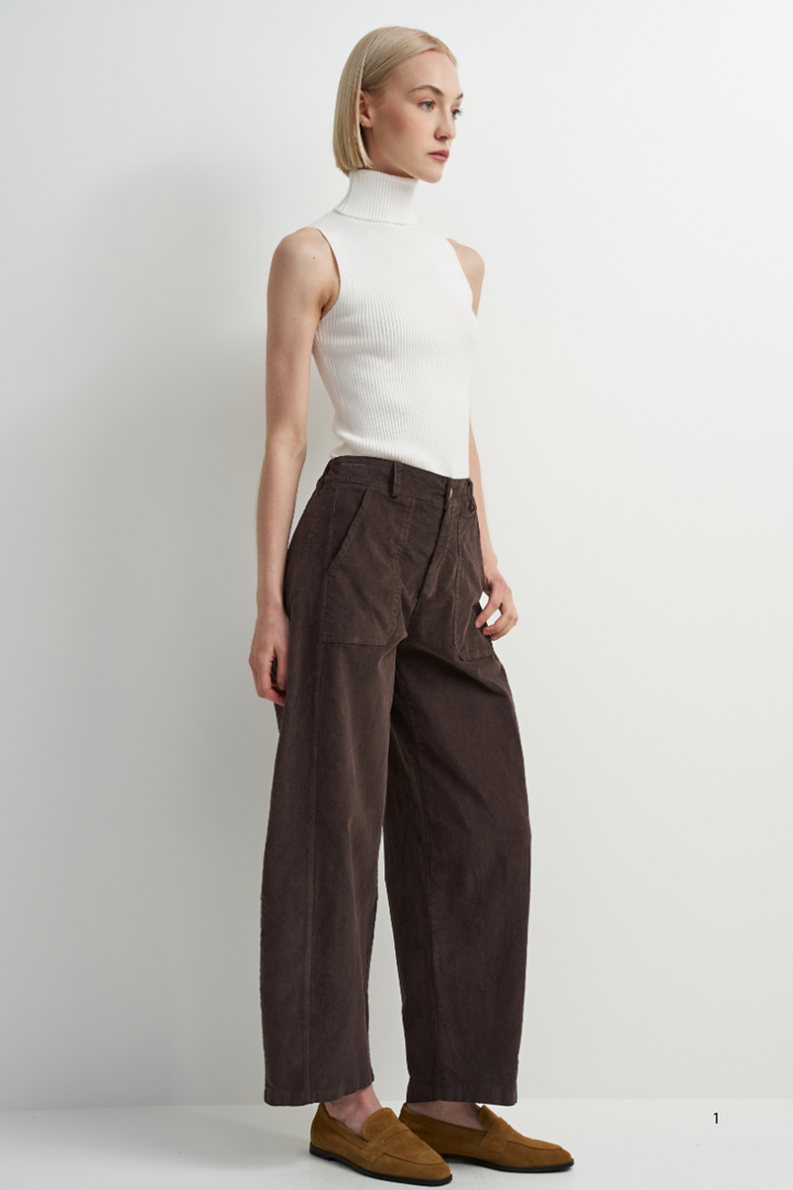 A model is shown from the side wearing a white sleeveless mock-neck top, dark brown wide-leg trousers, and brown suede loafers against a plain white studio background.
