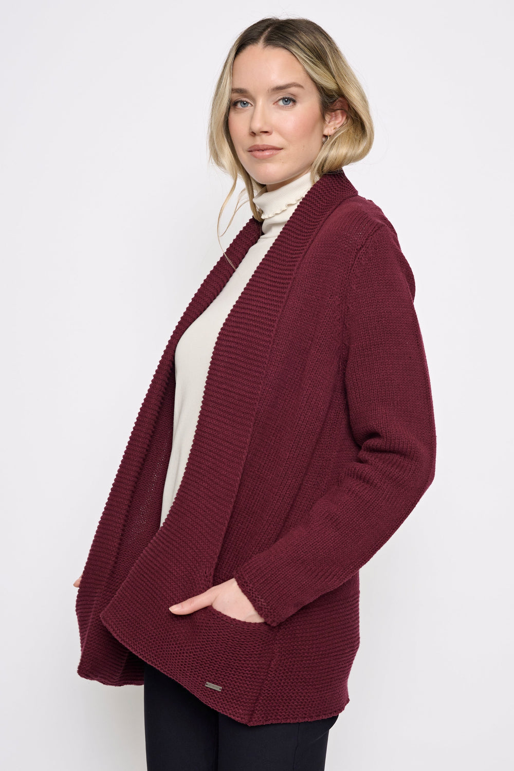A woman wearing a burgundy open-front ribbed knit cardigan over a cream turtleneck and black pants, standing against a plain light gray studio background.