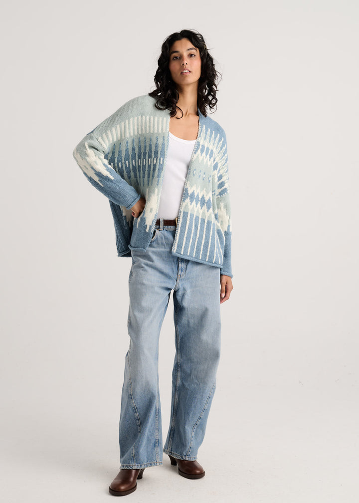 A woman modeling a light blue and white cardigan over a white tank top and light-wash blue jeans, standing against a plain light gray studio background.
