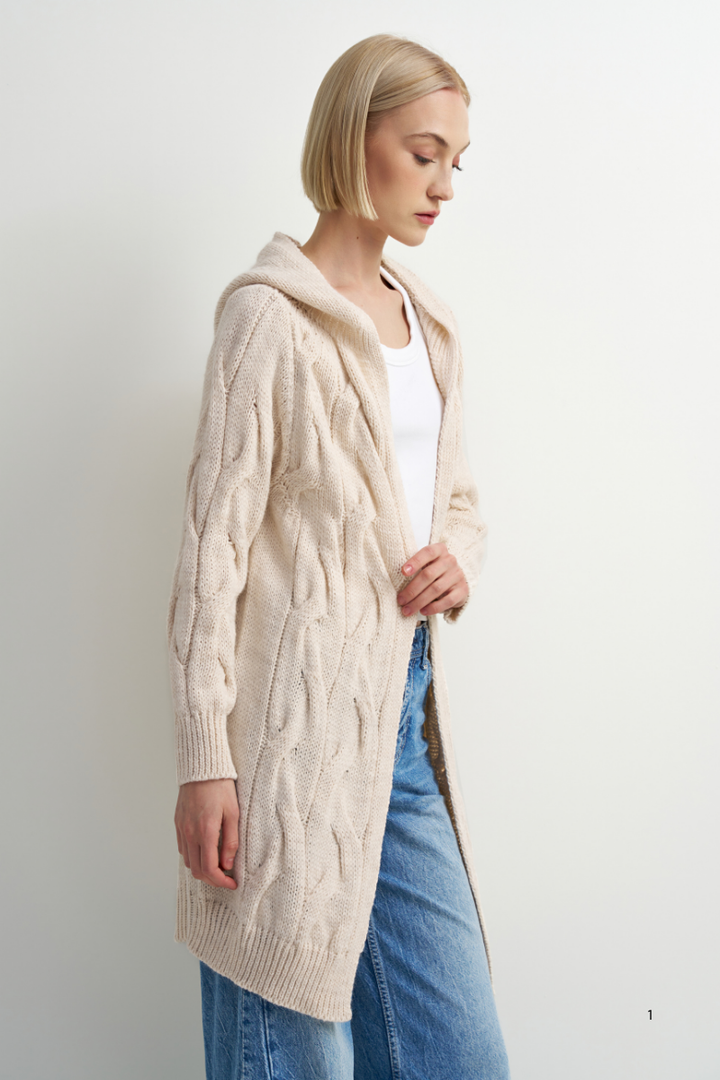 A woman wearing a long cream cable-knit hooded cardigan over a white top and blue jeans, standing against a plain light gray studio background.