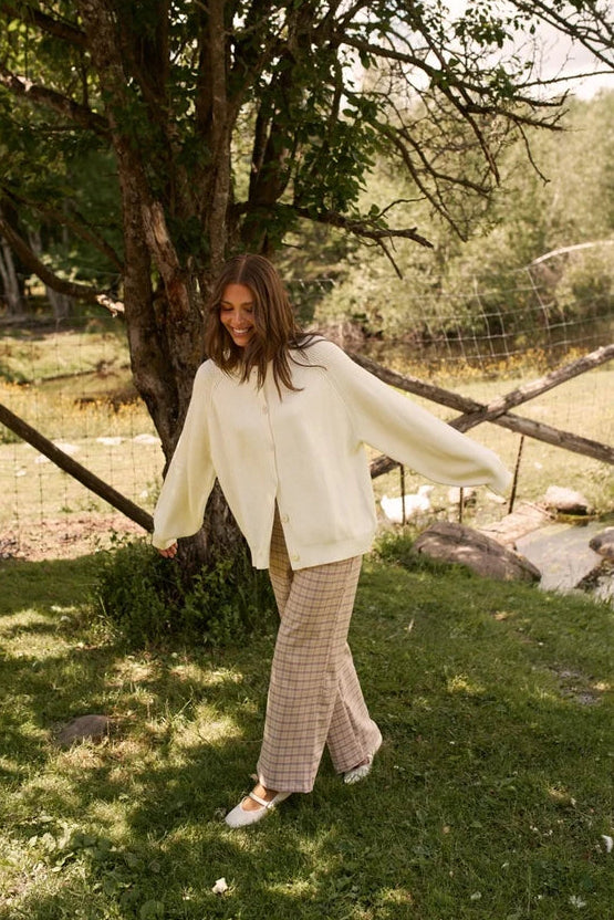 A woman stands outdoors on a grassy area near a tree, wearing a cream knit cardigan and beige plaid wide-leg pants.