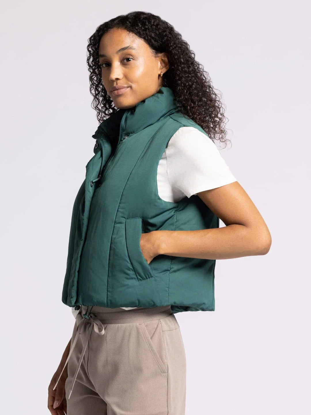 Woman wearing a green vest over a white shirt and beige shorts on a light gray background