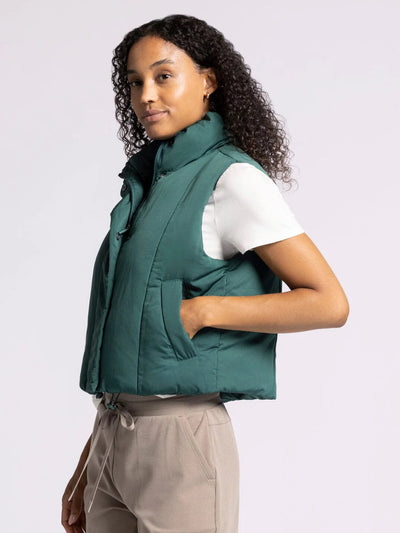 Woman wearing a green vest over a white shirt and beige shorts on a light gray background