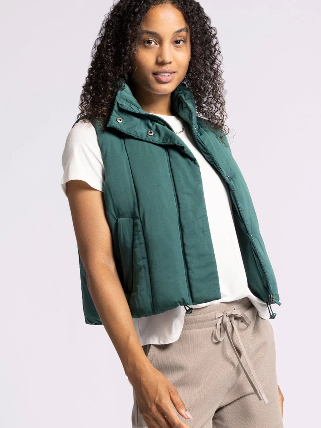 Person wearing a green puffer vest over a white shirt and beige pants on a light gray background