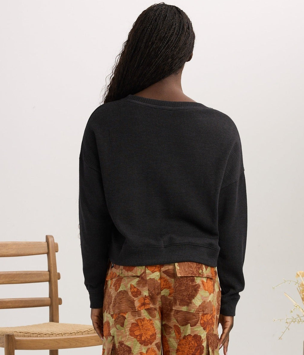 A model stands with their back to the camera wearing wide-leg trousers with a rust-orange and beige floral pattern, paired with a black sweatshirt. The setting is a studio with a wooden chair on the left and a patterned side table with a vase of dried grass on the right, all against a clean off-white background.