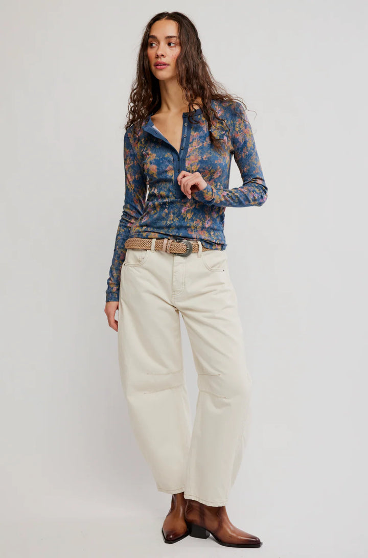 A woman modeling a long-sleeve button-up shirt with a blue and orange floral pattern, paired with cream wide-leg trousers and brown leather ankle boots, against a light gray studio background.