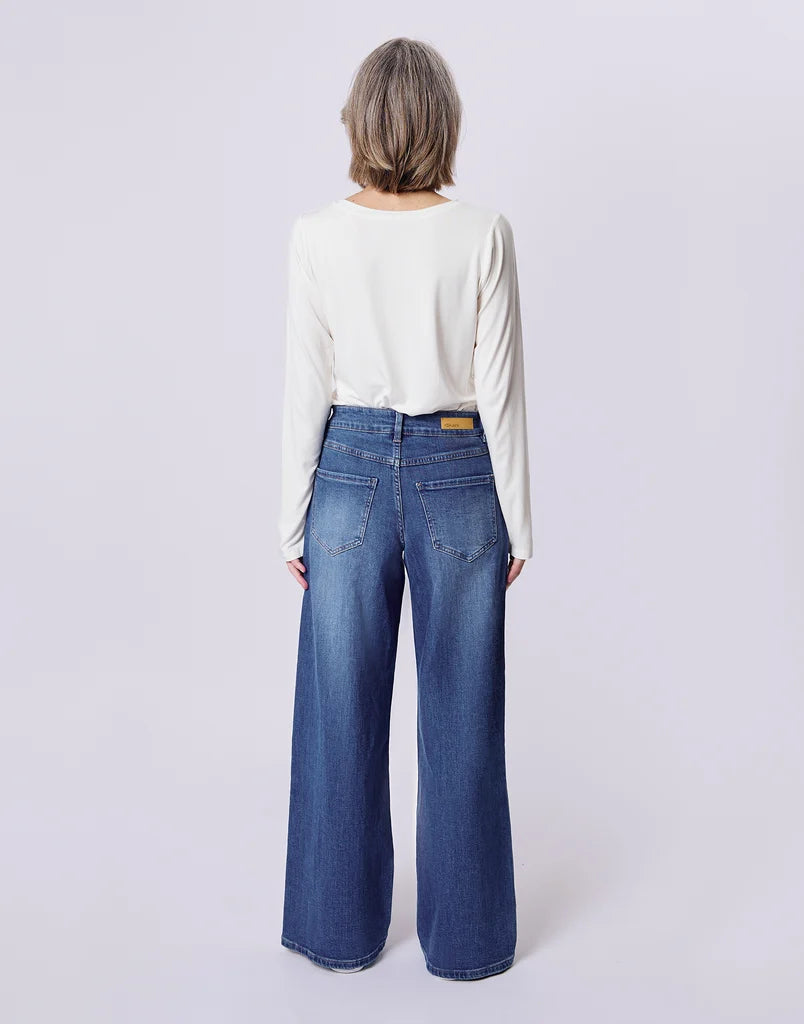 A model shown from the back wearing a white long-sleeve top and high-waisted, wide-leg blue denim jeans against a light gray studio background.