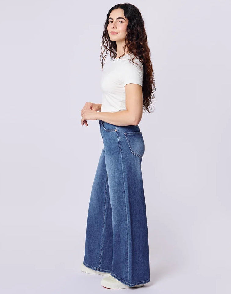 A woman modeling a pair of wide-leg blue jeans with a white short-sleeve t-shirt, standing against a light gray studio background.