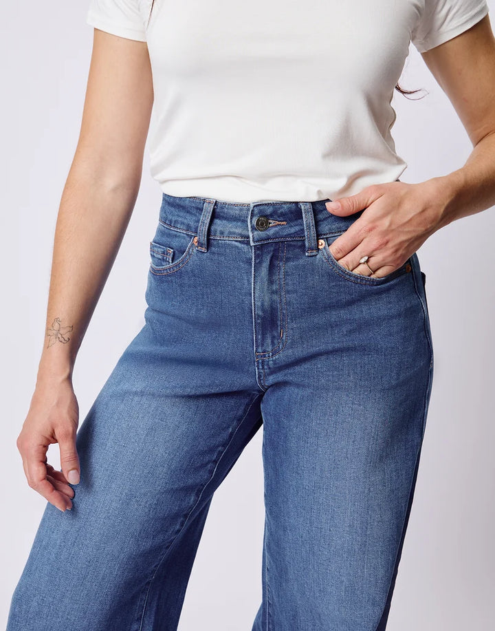 A person wearing blue denim jeans and a white short-sleeve top, cropped from the chest down, standing against a light gray studio background.