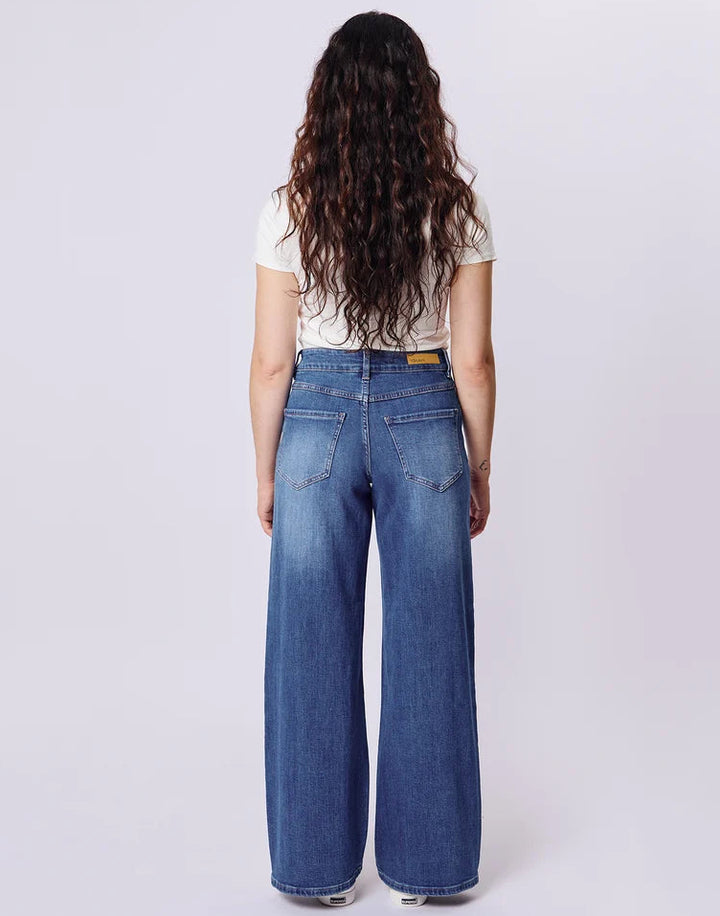 A woman viewed from behind wearing blue wide-leg denim jeans, a white short-sleeve t-shirt, and white sneakers against a light gray studio background.