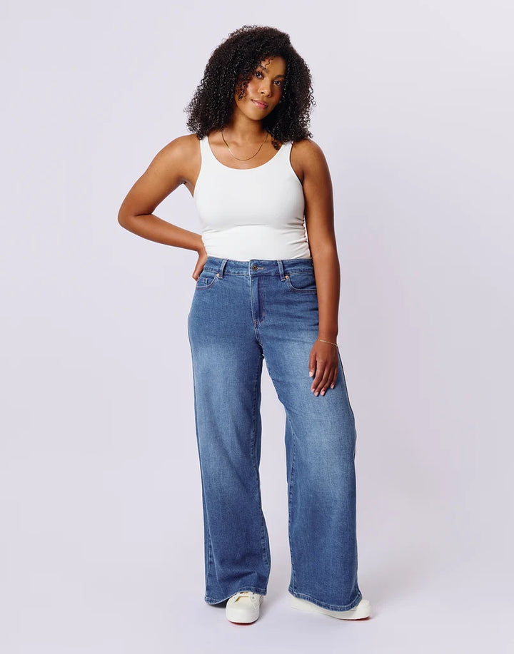 A woman modeling a pair of wide-leg blue jeans with a white tank top and white sneakers, standing against a light gray studio background.