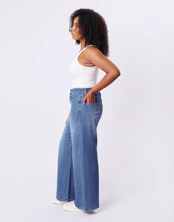 A woman shown in side profile wearing a white ribbed tank top and high-waisted, wide-leg blue denim jeans with white sneakers, set against a plain light gray studio background.
