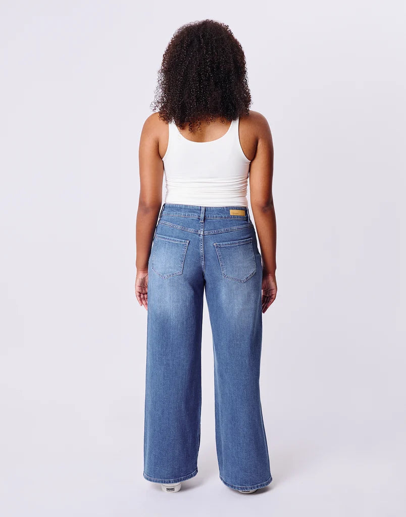 A model shown from the back wearing a pair of wide-leg blue jeans and a white ribbed tank top, standing against a light gray studio background.