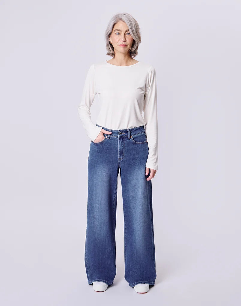 A woman with short gray hair stands against a light gray studio background wearing a cream long-sleeve top and blue wide-leg jeans.