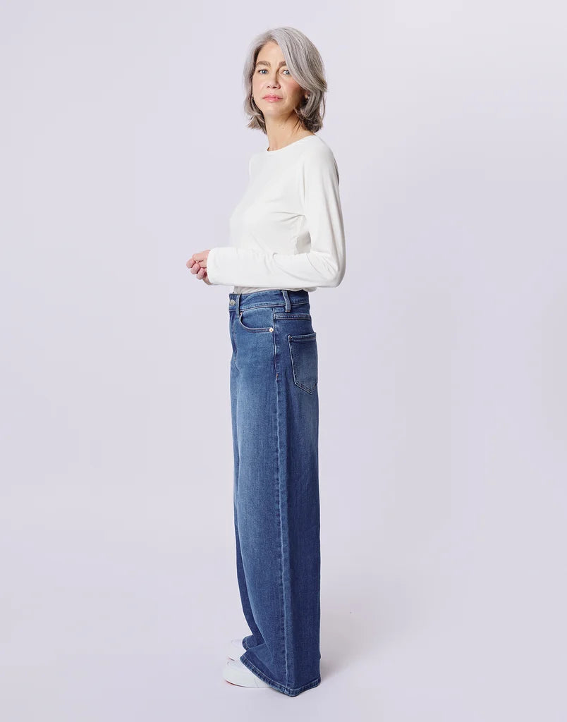 A woman with short gray hair stands in profile wearing a white long-sleeve top and high-waisted, wide-leg blue jeans against a light gray studio background.