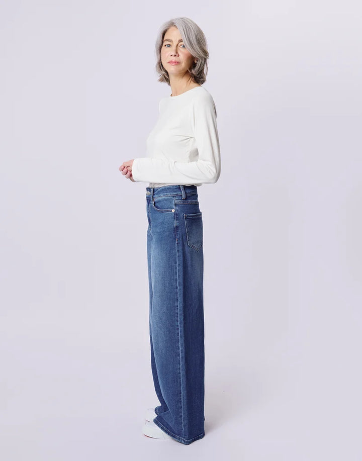 A woman with short gray hair stands in profile wearing a white long-sleeve top and high-waisted, wide-leg blue jeans against a light gray studio background.