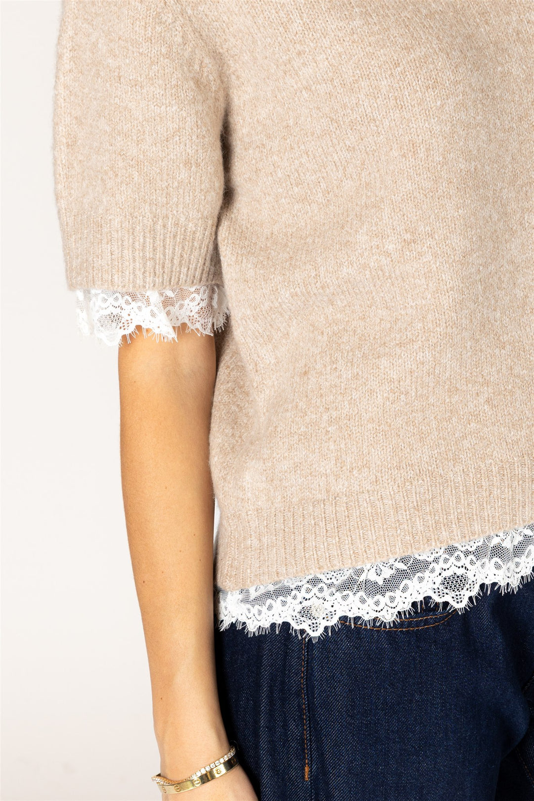 A close-up of a person wearing a beige knit sweater with white lace trim at the sleeve cuffs and hem, paired with dark blue jeans.