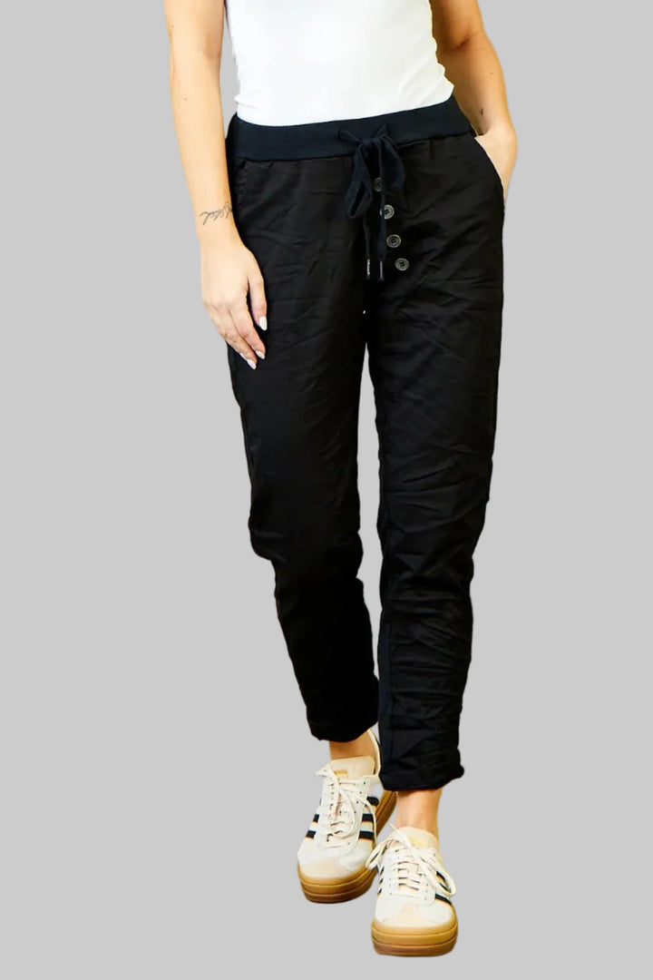 A woman modeling black drawstring pants with button details at the hips, paired with a white tank top and white sneakers, standing against a solid olive green studio background.