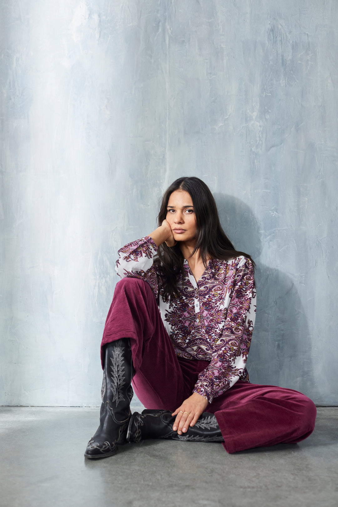 Woman sitting against a gray textured wall wearing a floral blouse and burgundy pants.