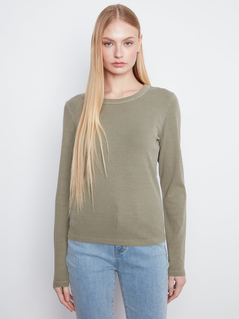 A blonde model wearing a long-sleeve crewneck knit sweater in olive green, paired with light blue jeans, against a plain light gray studio background.
